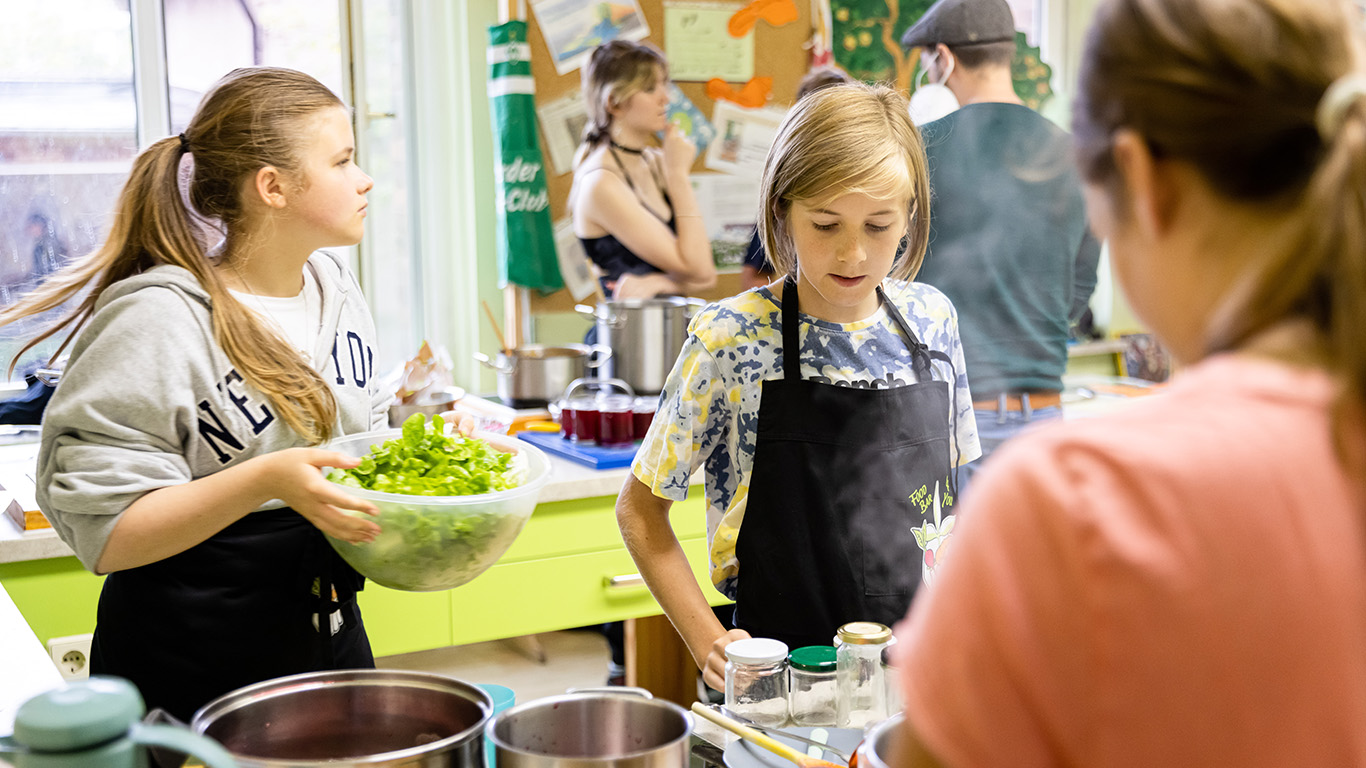 Kinder stehen in einer Küche und kochen. Ein Mädchen hält eine Schüssel Salat in der Hand.